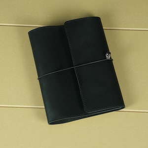 Pode incluir: Uma capa de couro preta para caderno com acabamento fosco liso, apresentando um fecho el&aacute;stico vertical envolvente preso por um pequeno e intrincado pingente de metal prateado posicionado na capa frontal para manter o caderno fechado.