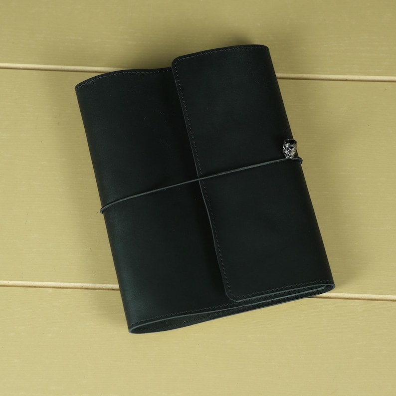 Pode incluir: Uma capa de couro preta para caderno com acabamento fosco liso, apresentando um fecho el&aacute;stico vertical envolvente preso por um pequeno e intrincado pingente de metal prateado posicionado na capa frontal para manter o caderno fechado.