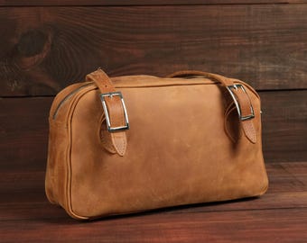 May include: A rectangular tan leather bag featuring a top zipper closure and two adjustable leather straps with silver-toned metal buckles attached to the front panel. The bag has a smooth, matte finish and a structured, horizontal orientation.