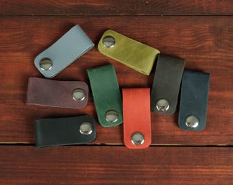 May include: A collection of eight rectangular leather key keepers in various colors including light blue, olive green, dark purple, forest green, black, orange, dark brown, and navy blue, each featuring a single silver-toned metal snap button closure.