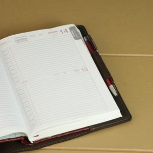 May include: An open daily planner with white pages featuring lined sections for time tracking, marked with the dates 14 and 15 in red text. A dark brown leather cover holds the pages, and a metallic red pen is secured in a side pen loop.