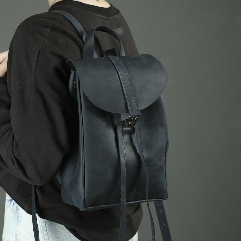 Pu&ograve; includere: Uno zaino in pelle blu navy scuro con chiusura a patta ripiegabile, dotata di un cinturino verticale e dettagli in metallo. La borsa presenta un manico superiore, spallacci regolabili e due lunghi e sottili lacci in pelle sul davanti.