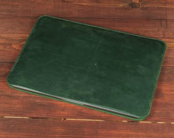 May include: A rectangular dark green leather laptop sleeve with visible stitching along the edges. The sleeve has a smooth, matte finish and a slim profile, designed to hold a flat electronic device securely within its protective, structured casing.