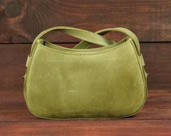 May include: A light olive green handbag with a smooth, matte finish and a curved top silhouette. The bag features a single integrated handle that arches over the top, visible stitching along the edges, and small metal hardware on the sides.