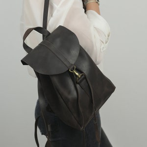 Pu&ograve; includere: Uno zaino in pelle marrone scuro caratterizzato da una chiusura a patta ripiegabile con gancio in metallo color ottone. La borsa include un manico superiore e spallacci regolabili, con una finitura liscia e opaca e un design strutturato.