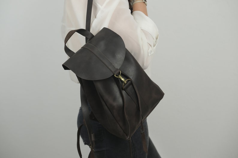 Pu&ograve; includere: Uno zaino in pelle marrone scuro caratterizzato da una chiusura a patta ripiegabile con gancio in metallo color ottone. La borsa include un manico superiore e spallacci regolabili, con una finitura liscia e opaca e un design strutturato.