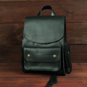 Può includere: Uno zaino nero caratterizzato da un ampio scomparto principale con chiusura a patta, una tasca frontale fissata da due bottoni a pressione in metallo color ottone e una maniglia superiore. La borsa ha un design strutturato con tasche laterali.