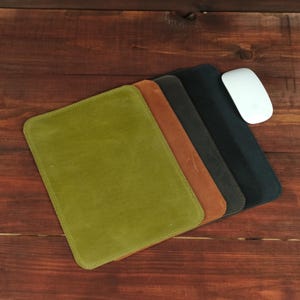 Op de afbeelding: Een set van vier rechthoekige lederen muismatten in olijfgroen, bruin, donkergrijs en zwart, gerangschikt in een verspringende stapel. Een witte computermuis rust aan de rechterkant van de zwarte muismat, wat de set accessoires compleet maakt.