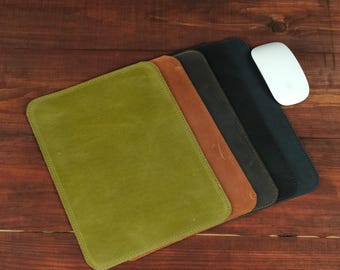 May include: A set of four rectangular leather mouse pads in olive green, tan, dark gray, and black, arranged in a staggered stack. A white computer mouse rests on the right side of the black mouse pad, completing the desk accessory set.