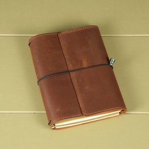 Pode incluir: Uma capa de couro castanho para caderno com um acabamento mate suave e marcas naturais vis&iacute;veis. A capa &eacute; fixada por um cord&atilde;o el&aacute;stico preto fino enrolado horizontalmente &agrave; volta do centro, com um pequeno pendente de metal prateado preso.