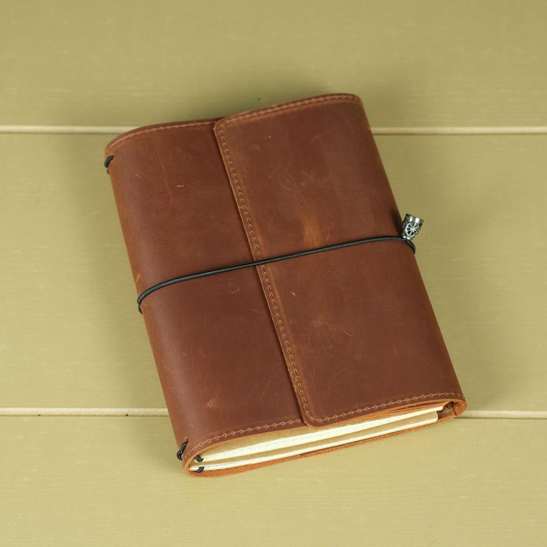 Pode incluir: Uma capa de couro castanho para caderno com um acabamento mate suave e marcas naturais vis&iacute;veis. A capa &eacute; fixada por um cord&atilde;o el&aacute;stico preto fino enrolado horizontalmente &agrave; volta do centro, com um pequeno pendente de metal prateado preso.