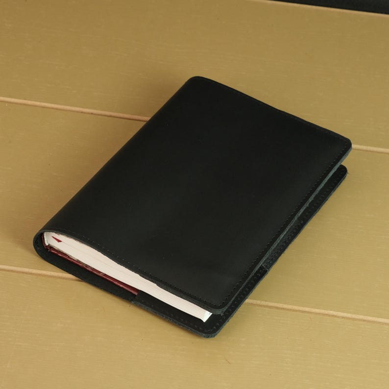 May include: A rectangular notebook featuring a smooth, solid black cover. The exterior is crafted from a dark, uniform material that wraps around the white pages visible along the side edge. The design is simple, functional, and clean.