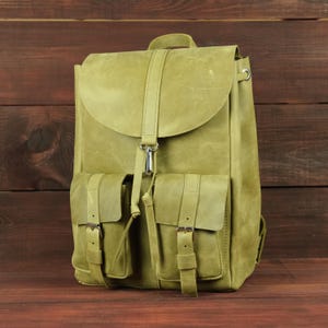 Può includere: Uno zaino in pelle verde oliva chiaro caratterizzato da una grande patta superiore con cinturino centrale verticale e chiusura con fibbia in metallo argentato. La parte anteriore include due tasche più piccole, ciascuna fissata con un cinturino in pelle e fibbia.