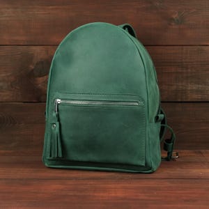 May include: A solid dark green backpack featuring a rounded top silhouette, a front zippered pocket with a silver-toned metal zipper, and a matching leather tassel pull attached to the zipper slider. The bag has adjustable shoulder straps.