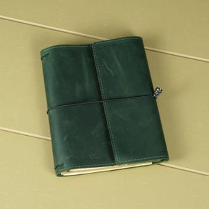 Pode incluir: Uma capa de di&aacute;rio em couro verde-escuro com acabamento fosco e textura de gr&atilde;o natural vis&iacute;vel. A capa &eacute; presa por um cord&atilde;o el&aacute;stico preto fino enrolado horizontalmente ao redor do centro, com um pequeno pingente de metal prateado anexado.