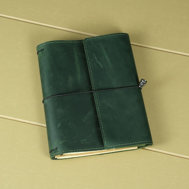 Pode incluir: Uma capa de di&aacute;rio em couro verde-escuro com acabamento fosco e textura de gr&atilde;o natural vis&iacute;vel. A capa &eacute; presa por um cord&atilde;o el&aacute;stico preto fino enrolado horizontalmente ao redor do centro, com um pequeno pingente de metal prateado anexado.