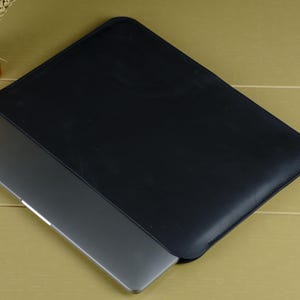 May include: A dark gray laptop partially inserted into a black rectangular sleeve. The sleeve features rounded corners and visible stitching along the edges, providing a snug fit for the device. The laptop has a smooth, metallic finish.