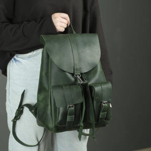 Può includere: Uno zaino in pelle verde bosco scuro caratterizzato da una grande patta ripiegabile con chiusura centrale in metallo, due tasche frontali con cinturini con fibbia regolabili e spallacci in pelle verde abbinati con finiture e rivetti in metallo.