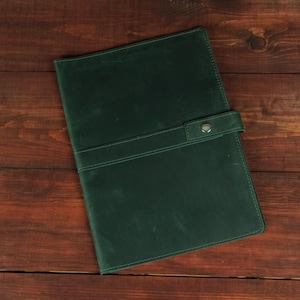 Può includere: Una copertina per taccuino in pelle verde scuro con una finitura opaca liscia e una cinghia di chiusura orizzontale fissata con un bottone a pressione in metallo color argento. La copertina rettangolare ha angoli arrotondati e cuciture.
