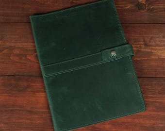 May include: A dark green leather notebook cover featuring a smooth matte finish and a horizontal strap closure secured with a single silver-toned metal snap button. The rectangular cover has rounded corners and visible stitching along the edges.