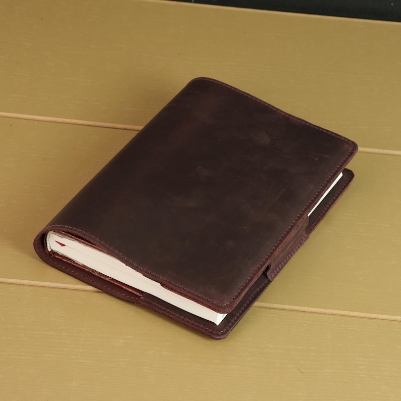 May include: A dark brown leather notebook cover with a smooth, matte finish and visible stitching along the edges. The cover is wrapped securely around a thick white paper notebook, featuring a rectangular flap closure on the right side.