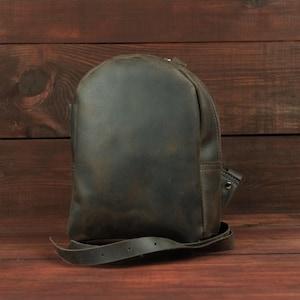 Könnte beinhalten: Ein dunkelolivgrüner Lederrucksack mit glatter, matter Oberfläche. Die Tasche hat eine abgerundete Oberseite, ein einzelnes Hauptfach und einen langen, verstellbaren Lederriemen mit sichtbaren Metallnieten am seitlichen Befestigungspunkt.