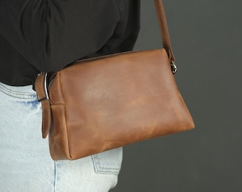 May include: A rectangular brown leather crossbody bag with a smooth finish, featuring a top zipper closure, a matching adjustable leather shoulder strap, and silver-toned metal hardware attachments on the sides of the bag.