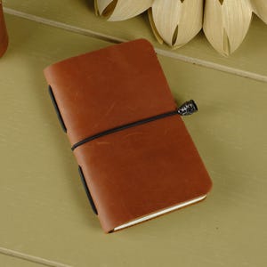 May include: A rectangular notebook with a smooth, tan-colored leather cover. The notebook is secured by a thin black elastic band that wraps horizontally around the center, featuring a small, dark, decorative metal bead on the right side.