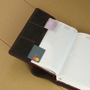 May include: A dark brown leather notebook cover featuring multiple interior slots for organization. The cover holds an open planner with lined pages, a small rectangular paper slip, and a dark gray payment card with a visible red and yellow logo.