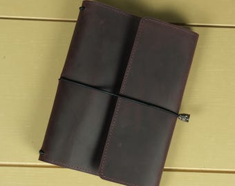 May include: A dark burgundy leather journal cover featuring a smooth matte finish and rounded corners. The cover is secured by a thin black elastic cord that wraps horizontally around the center, finished with a small metallic charm at the end.