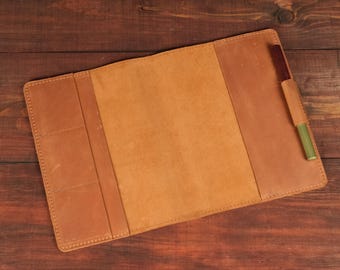 May include: A tan leather notebook cover featuring a central panel, two side flaps with stitched edges, and a dedicated pen loop on the right side holding a metallic pen with a dark brown cap and a gold-toned body.