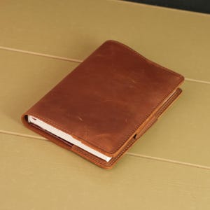 May include: A rectangular notebook encased in a smooth, matte brown leather cover. The leather features visible natural grain and subtle surface variations. The white pages of the notebook are partially visible along the side edge of the cover.