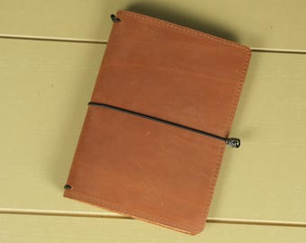 May include: A rectangular notebook cover made of smooth, tan-colored leather. The cover features a single black elastic band wrapped horizontally around the center to keep it closed, with visible stitching along the edges of the leather material.
