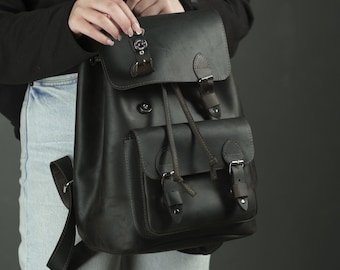 May include: A matte black leather backpack featuring a large main compartment with a fold-over flap, a front pocket with a flap, dark brown leather straps with silver-toned metal buckles, and a drawstring closure for the main storage area.