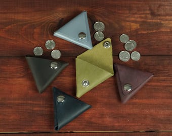 May include: Five triangular coin pouches in light blue, dark gray, olive green, navy blue, and deep purple. Each pouch features a single silver-toned metal snap button closure. Several small silver-colored coins are scattered around the pouches.