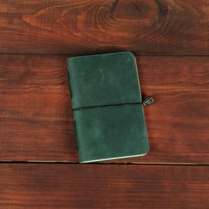May include: A dark green leather journal with a smooth, matte finish and rounded corners. The notebook is secured by a thin black elastic band that wraps horizontally around the cover, featuring a small, decorative metallic charm attached to the side.