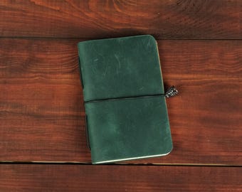 May include: A dark green leather journal with a smooth, matte finish and rounded corners. The notebook is secured by a thin black elastic band that wraps horizontally around the cover, featuring a small, decorative metallic charm attached to the side.