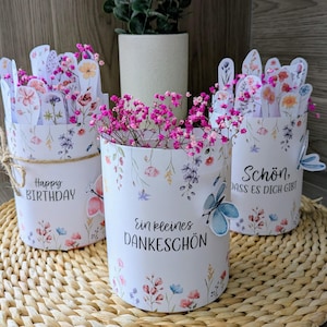 Puede incluir: Tres recipientes cilíndricos blancos con estampado floral llenos de flores rosas, cada uno con un mensaje diferente en alemán. Una pequeña planta en maceta está en el fondo. Los recipientes están sobre una estera tejida.