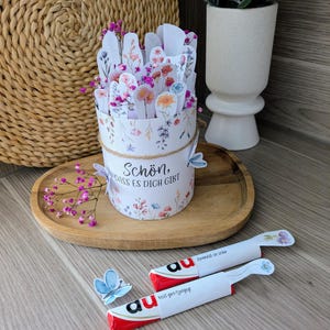 May include: A floral-themed gift box with the text "Sch&ouml;n, dass es dich gibt" and a decorative butterfly. The box is filled with paper flowers and sits on a wooden tray. Two wrapped chocolate bars are also present.
