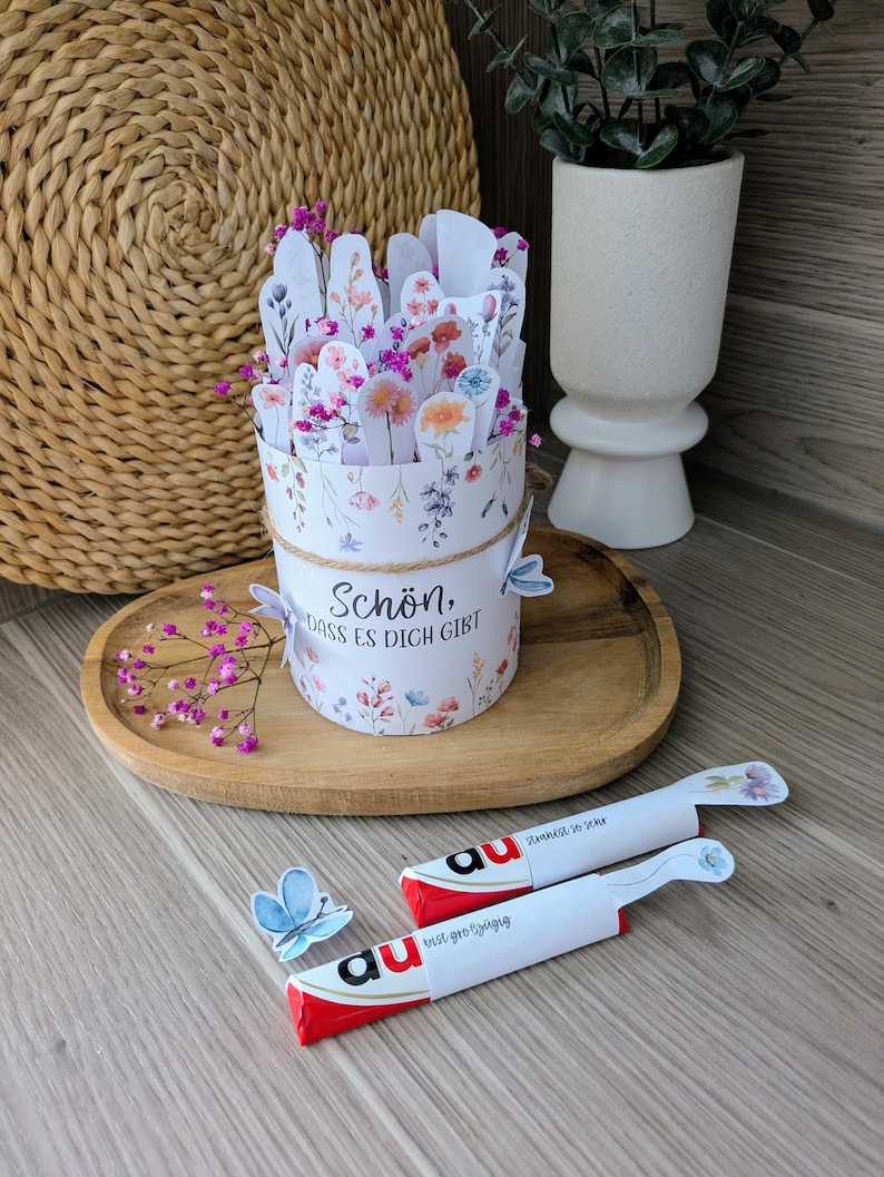 May include: A floral-themed gift box with the text "Sch&ouml;n, dass es dich gibt" and a decorative butterfly. The box is filled with paper flowers and sits on a wooden tray. Two wrapped chocolate bars are also present.