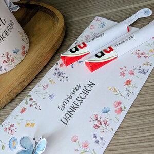 May include: Two white wrapped candies with red and black accents, resting on a floral card with the words "Ein kleines DANKESCH&Ouml;N". A blue butterfly sticker and a floral design are also present.