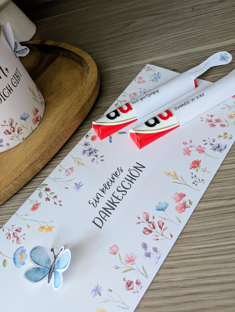 May include: Two white wrapped candies with red and black accents, resting on a floral card with the words "Ein kleines DANKESCH&Ouml;N". A blue butterfly sticker and a floral design are also present.