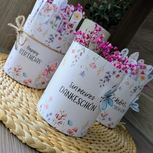 May include: Three white paper flower pots with floral designs and text. One pot says "Happy Birthday" and is tied with twine. The other pots say "Ein kleines Dankesch&ouml;n" and "Sch&ouml;n, dich gibt". Each pot is filled with pink flowers.