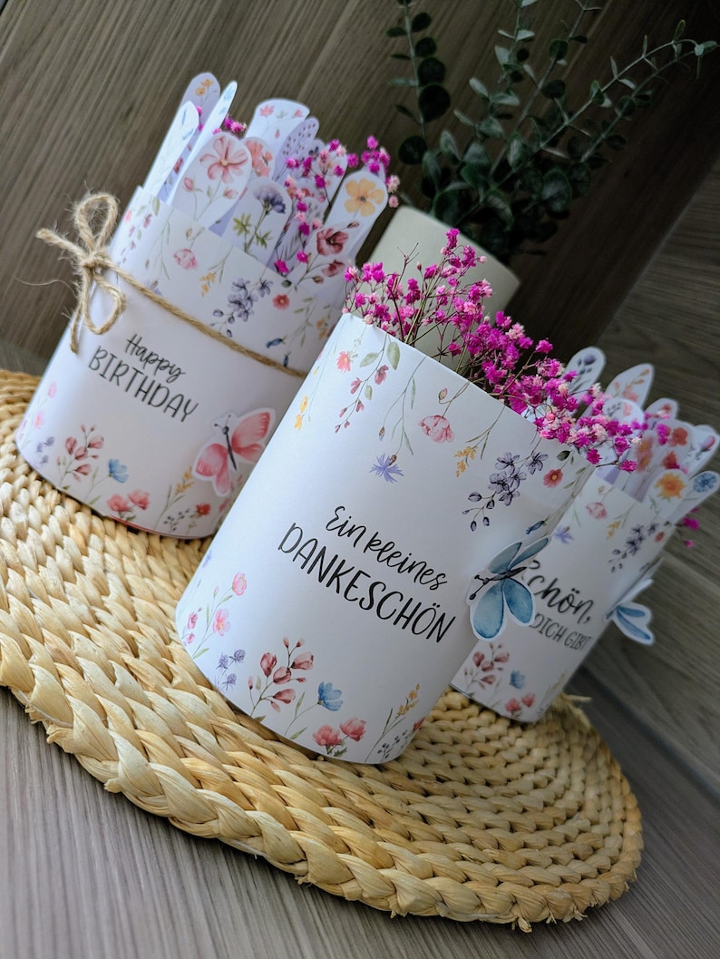 May include: Three white paper flower pots with floral designs and text. One pot says "Happy Birthday" and is tied with twine. The other pots say "Ein kleines Dankesch&ouml;n" and "Sch&ouml;n, dich gibt". Each pot is filled with pink flowers.