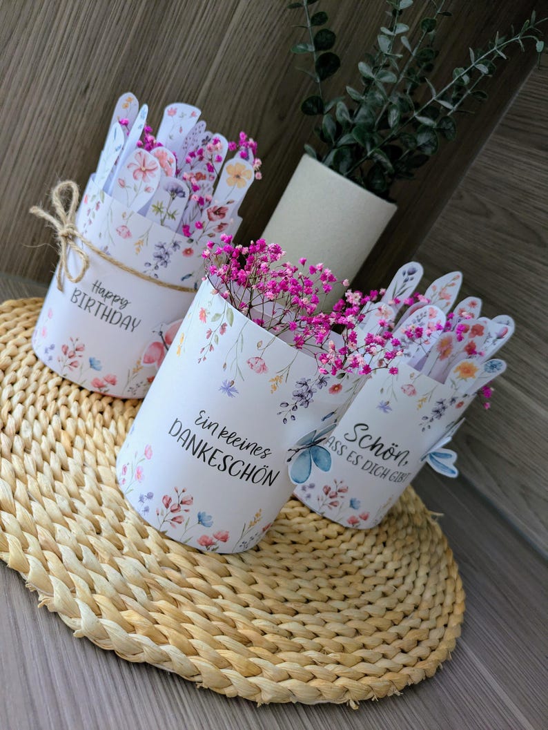May include: Three white floral-patterned paper vases with pink flowers. One vase says "Happy Birthday" and is tied with twine. The other two vases have German text. The vases sit on a woven mat.