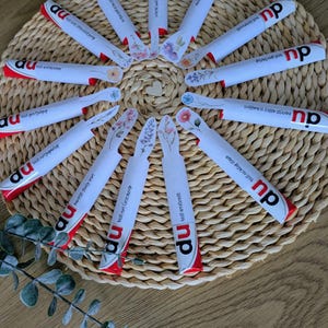 May include: A collection of white and red wrapped products arranged in a circular pattern on a woven mat. Each product features floral designs and text. The products are arranged around a small wooden heart.