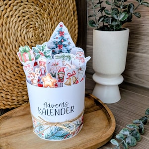 Könnte beinhalten: Ein weißer, zylindrischer Adventskalender mit festlichem Weihnachtsdesign. Der Kalender ist mit Illustrationen eines Weihnachtsbaums, Geschenken und Schneemännern verziert. Der Text "Advents KALENDER" ist auf der Vorderseite aufgedruckt und mit Bindfaden gebunden.