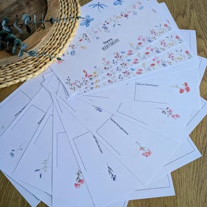 May include: A stack of white greeting cards with floral designs and the words "Happy Birthday." The cards feature watercolor illustrations of flowers, dragonflies, and botanical elements. Some cards have lines for writing.
