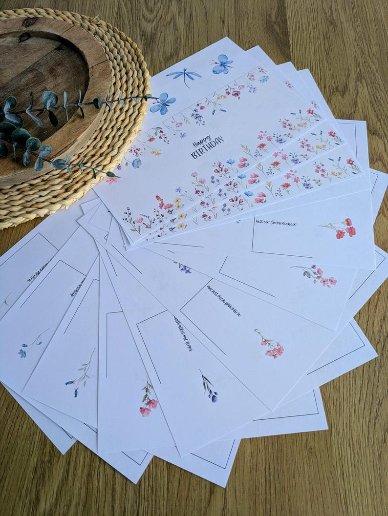 May include: A stack of white greeting cards with floral designs and the words "Happy Birthday." The cards feature watercolor illustrations of flowers, dragonflies, and botanical elements. Some cards have lines for writing.