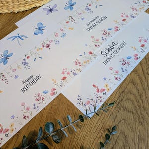 May include: White greeting cards with watercolor floral and insect designs. The cards feature dragonflies, butterflies, and floral borders in pink, blue, and yellow. Text includes "Happy Birthday" and other phrases.